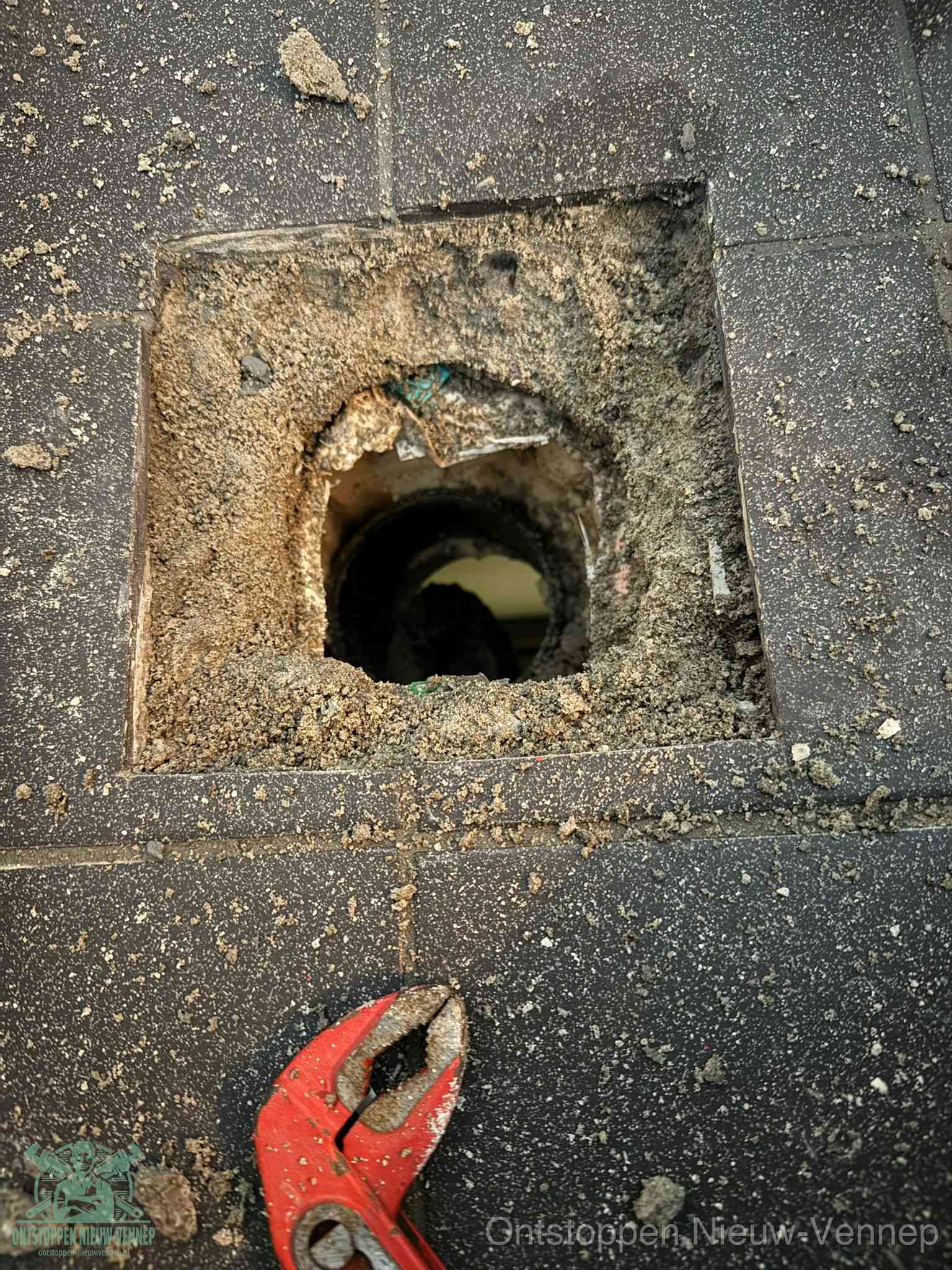 Een geopend rioolputje in een betontegelvloer met losgemaakt rooster en een zichtbare ronde afvoerbuis, het achterhalen waar de afvoerverstopping zit in Nieuw-Vennep