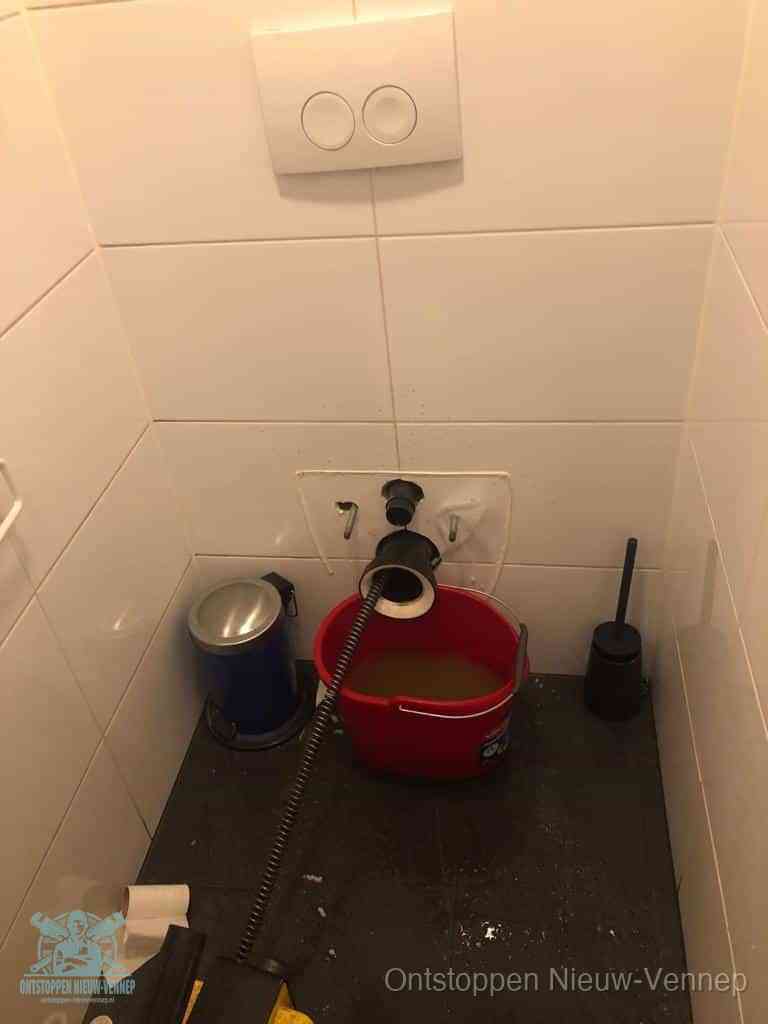 Een ontstopte wand-wc met verwijderde toiletpot waar een ontstoppingsveer in de wanduitgang verdwijnt, het achterhalen waar de verstopping zit in Nieuw-Vennep