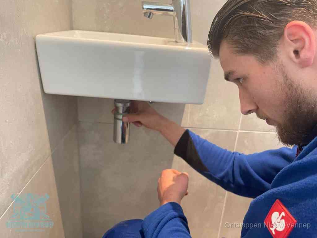 Een monteur draait met de hand de zwanenhalssifon onder een fonteinwasbak in de toiletruimte los om de bron van rioollucht in Nieuw-Vennep te vinden
