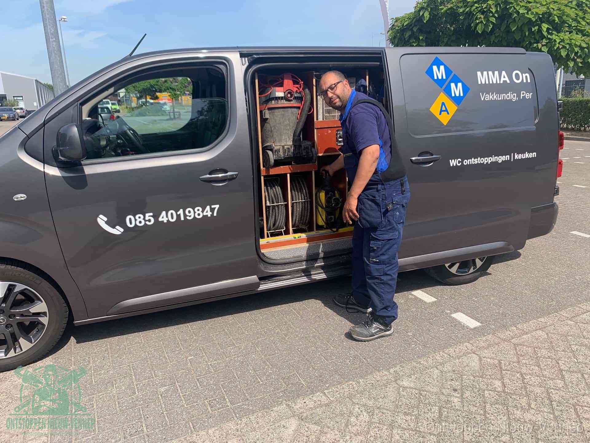Een monteur bij de open schuifdeur van zijn bedrijfsbus op een parkeerplaats, klaar om de oorzaak van terugkerende afvoerverstoppingen aan te pakken in Nieuw-Vennep