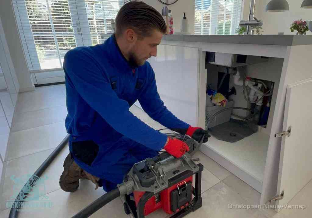 Een monteur voert een professionele elektrische ontstoppingsmachine in de afvoer onder de spoelbak tijdens het gootsteen ontstoppen in Nieuw-Vennep